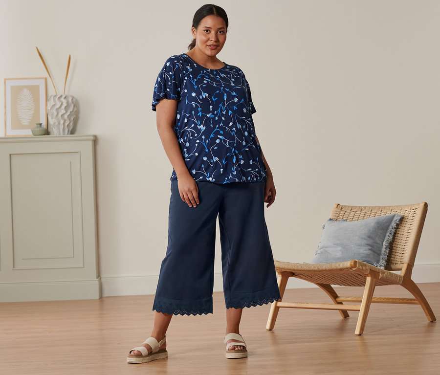 Žena stojí a má na sobě tmavě modré halenkové triko s netopýřími rukávy, culottes s výšivkou a kožené sandály na platformě.