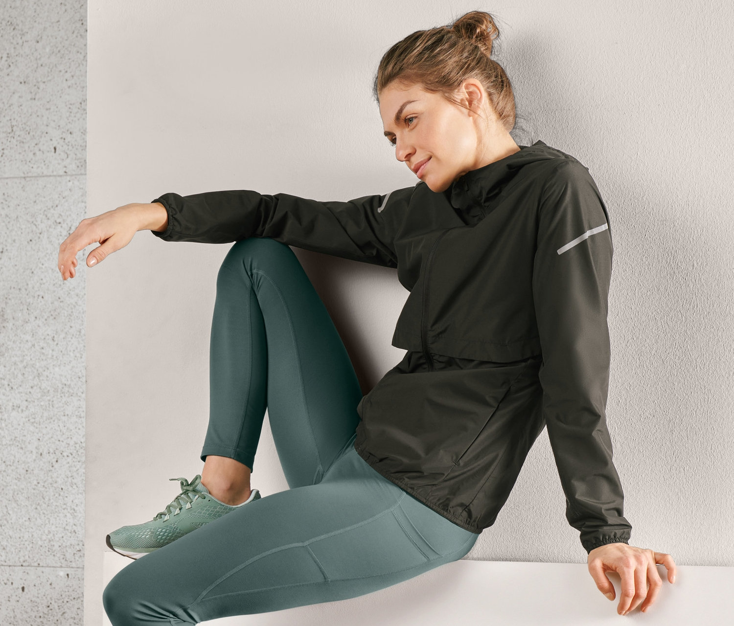Žena sedí na okraji a má na sobě černou větruvzdornou bundu na běhání, zelené legíny a tenisky.
