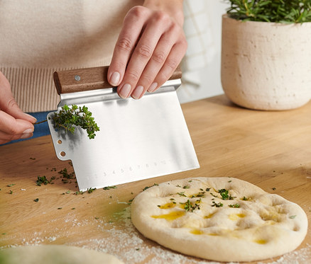 Žena krájí bylinky pomocí Kráječe na těsto 2 v 1 na dřevěném stole. Na stole leží těsto, olivový olej a rostlina v květináči.