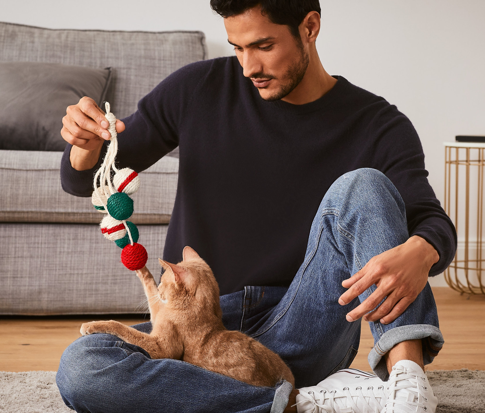 Muž sedí na podlaze a hraje si s kočkou s hračkou pro kočky »Chrastítko«.