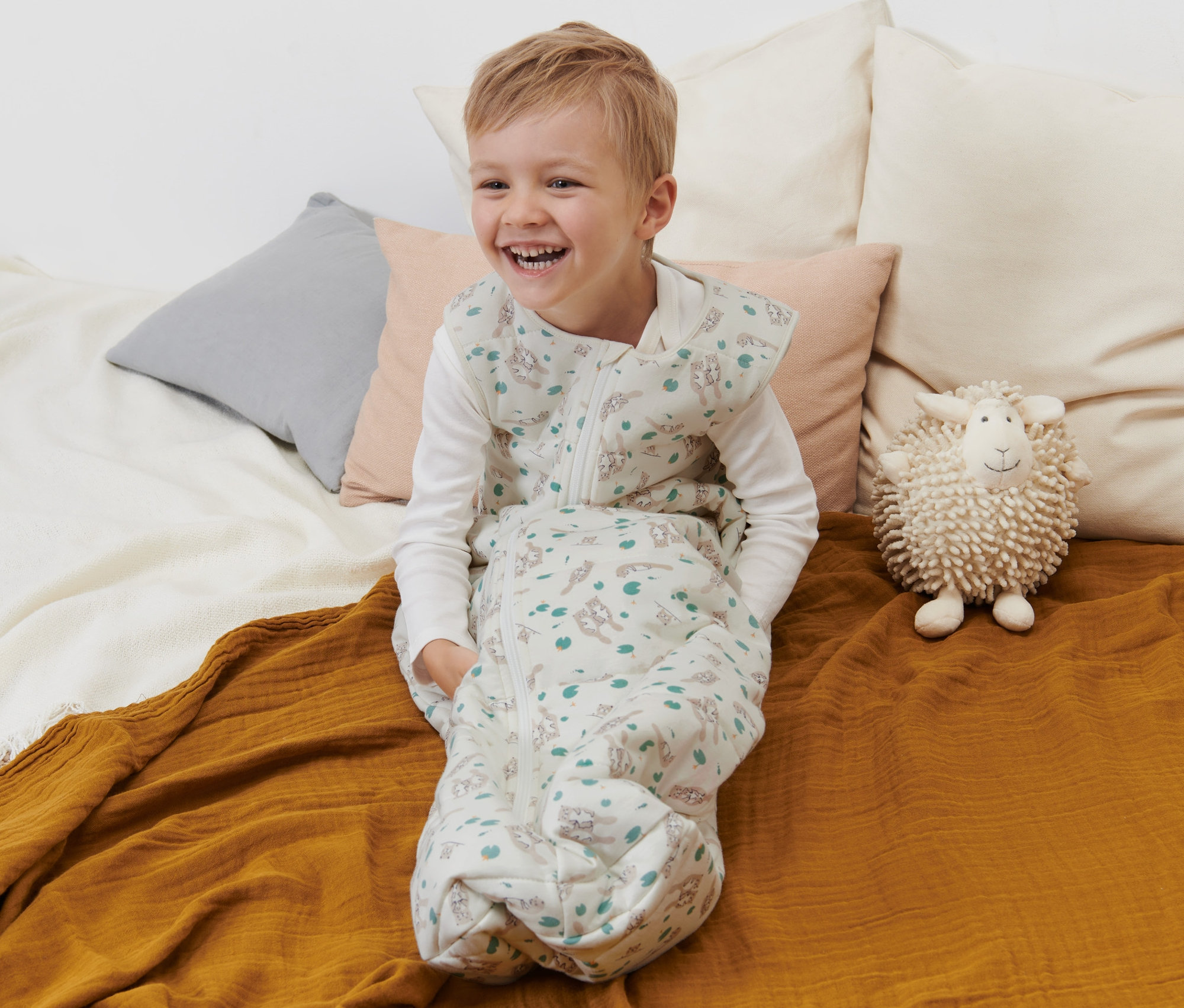 Usmívající se chlapec sedí v spacím pytli 2,5 TOG, 110 cm, s potiskem vyder na posteli.