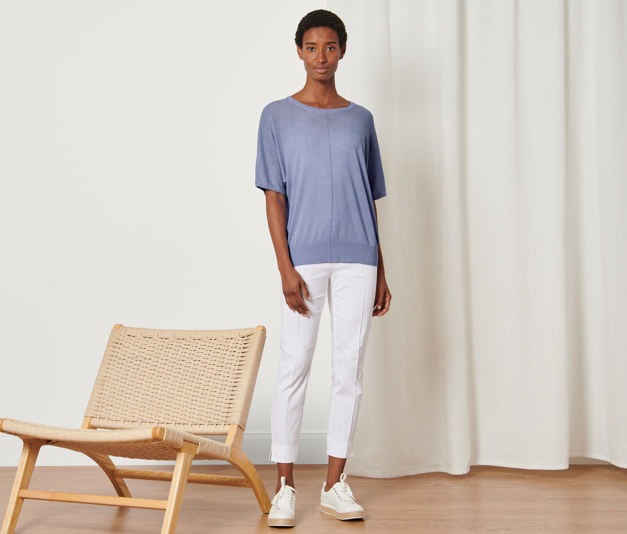 Žena stojí vedle proutěného křesla a má na sobě tričko z jemného úpletu z lněné směsi, bílé kalhoty a tenisky.