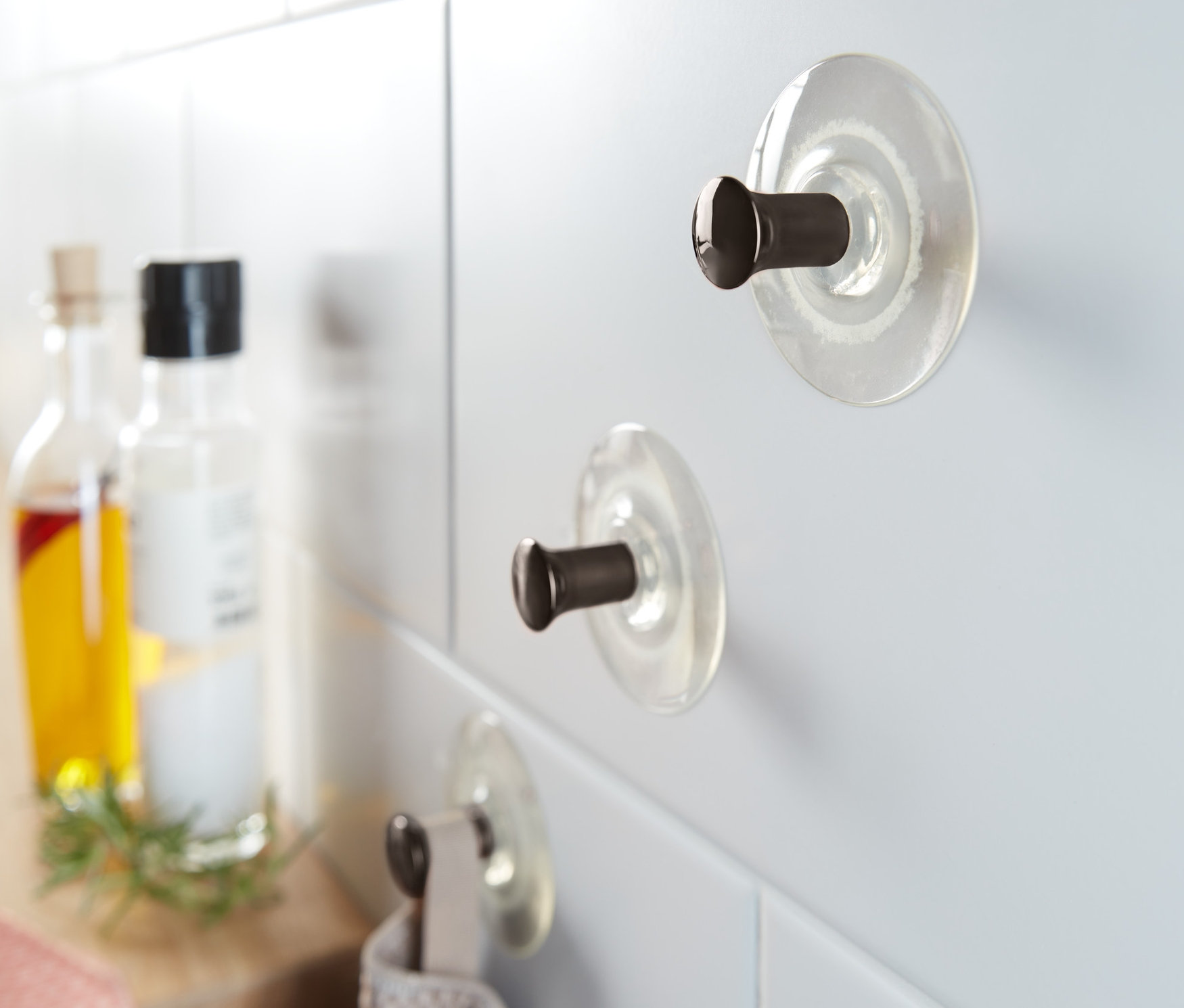 Na bílých dlaždicích jsou připevněny 3 ks háčků s přísavkou. Na jednom z nich visí šedý ručník.