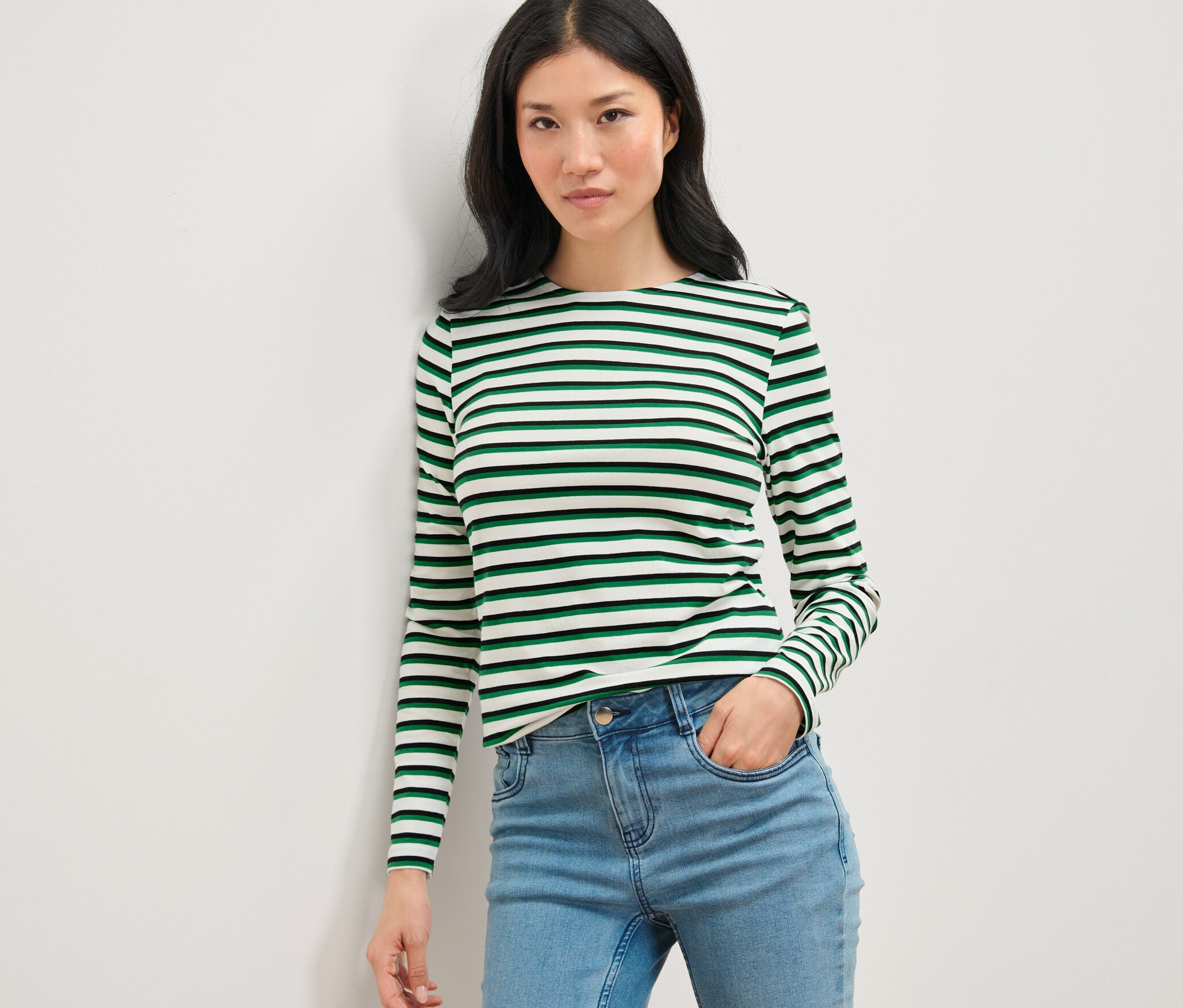 Žena pózuje v pruhovaném tričku s dlouhým rukávem v různých barvách a v džínách.