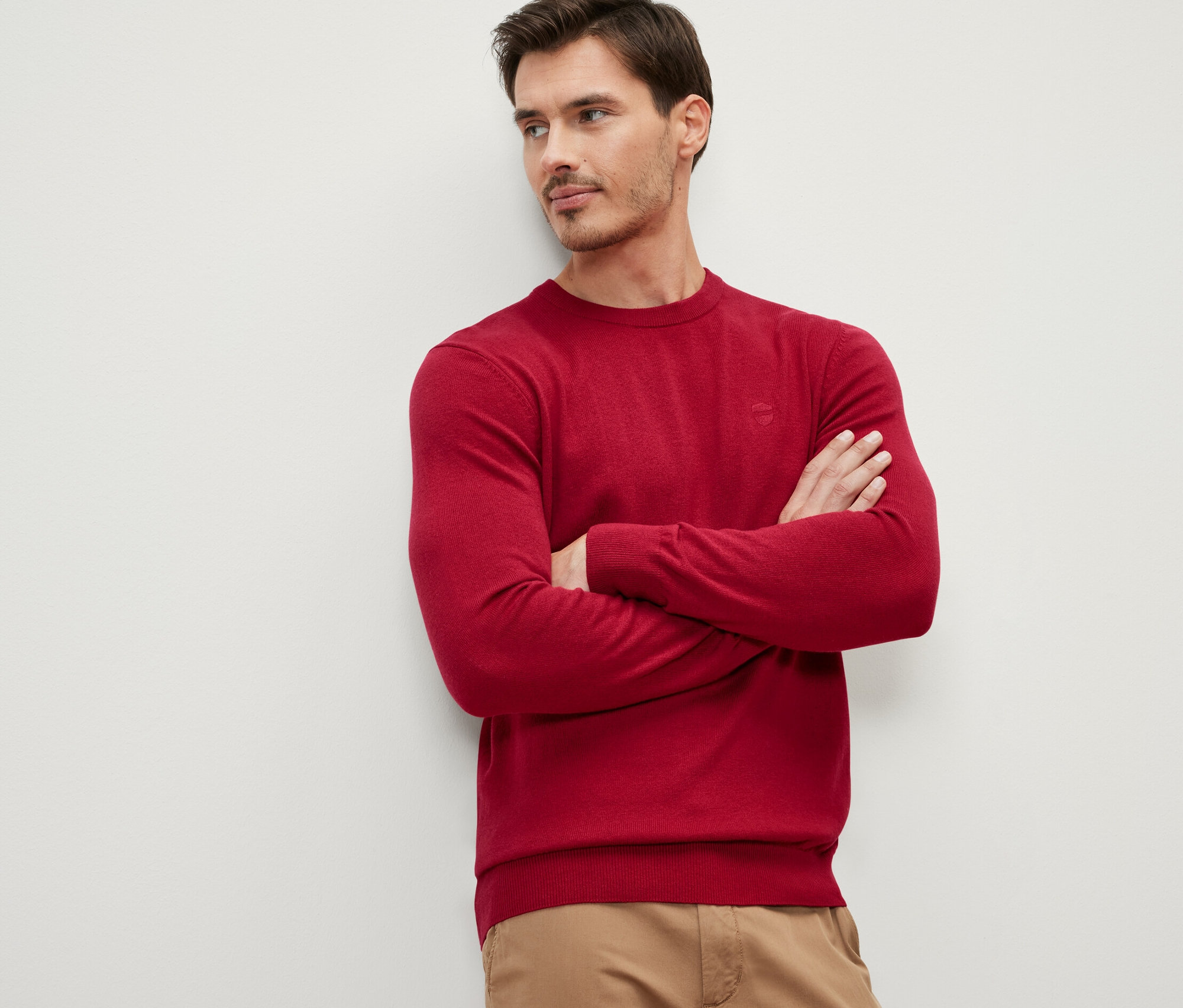 Muž se zkříženýma rukama na hrudi má na sobě červený svetr s kulatým výstřihem.