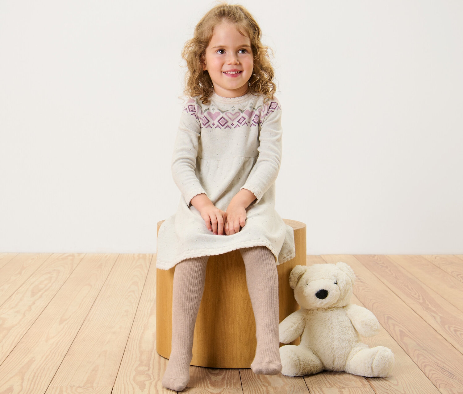Dívka sedí na dřevěné stoličce a má na sobě šaty a dětské punčocháče s vlnou RWS.