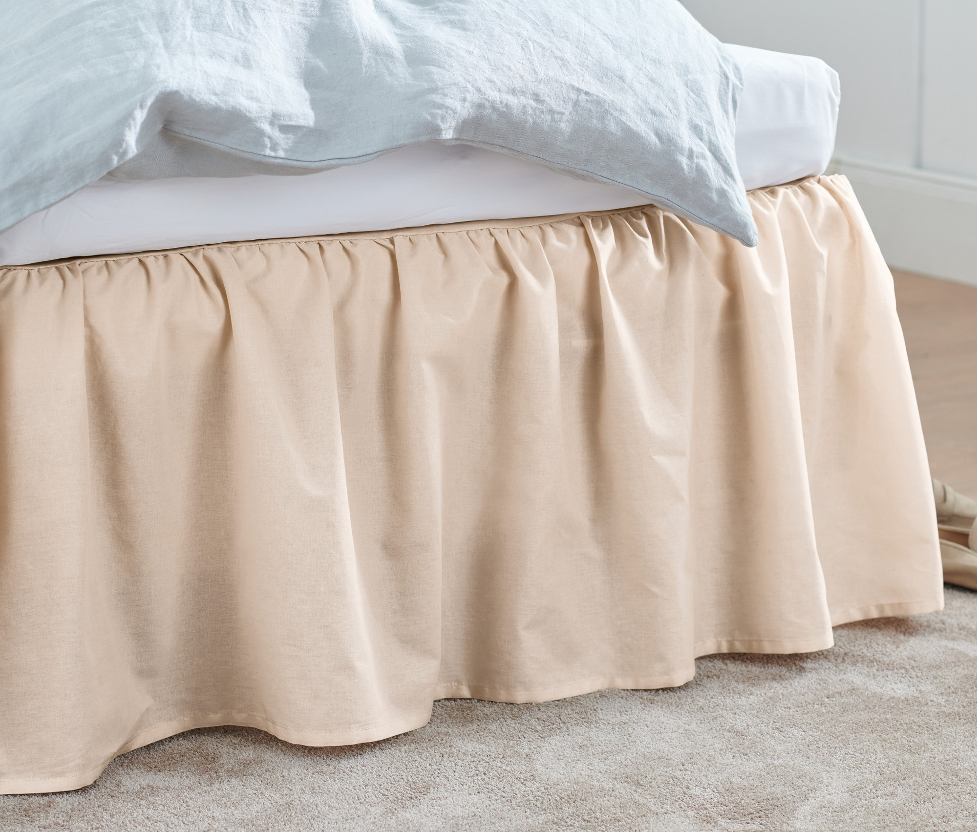 Béžový přehoz na postel zakrývá postel. U podlahy leží béžové boty a šedý koberec.