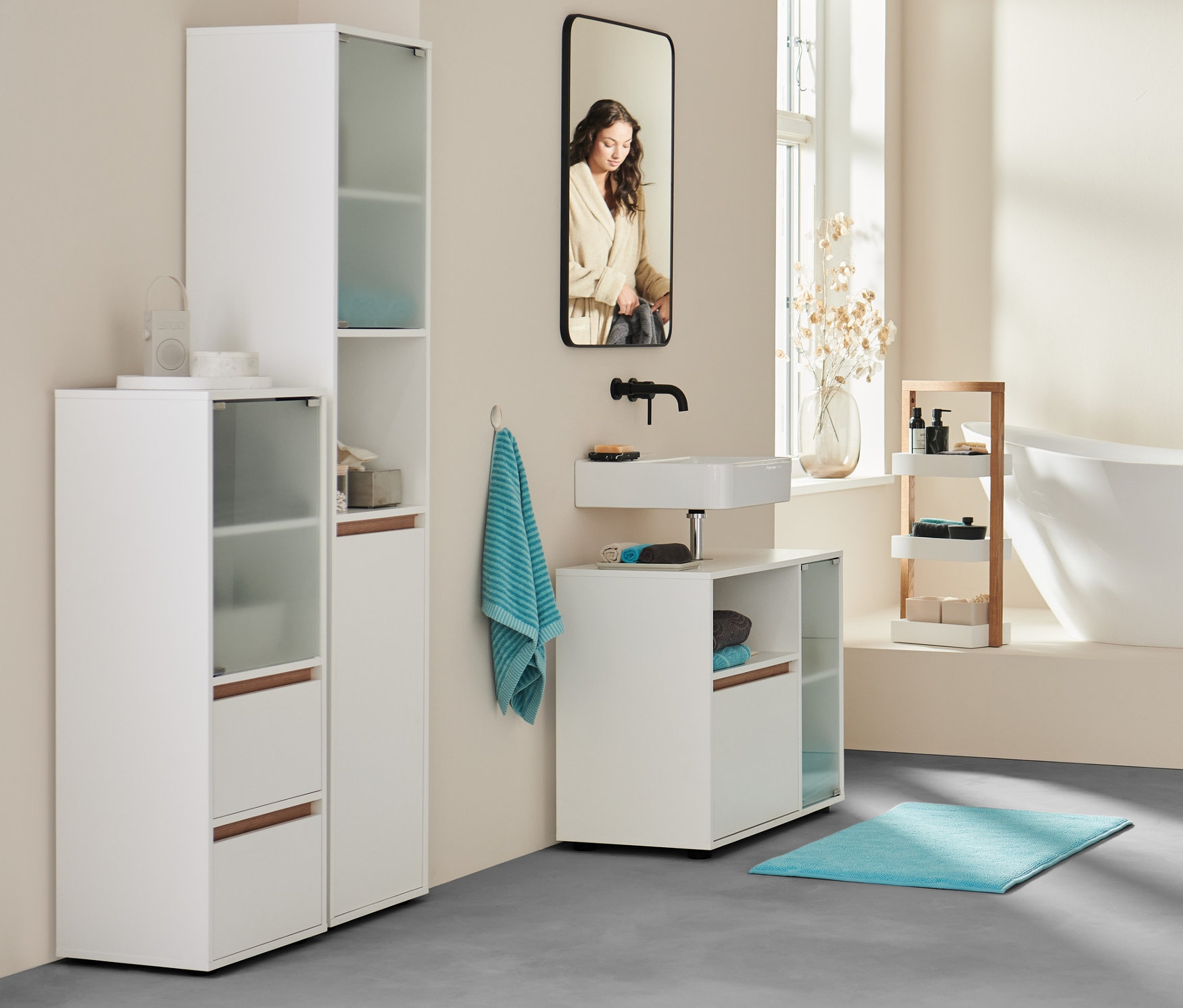 V koupelně stojí žena v županu u vany. V popředí koupelnová komoda, extra velká osuška antracitové barvy a modrozelené ručníky.