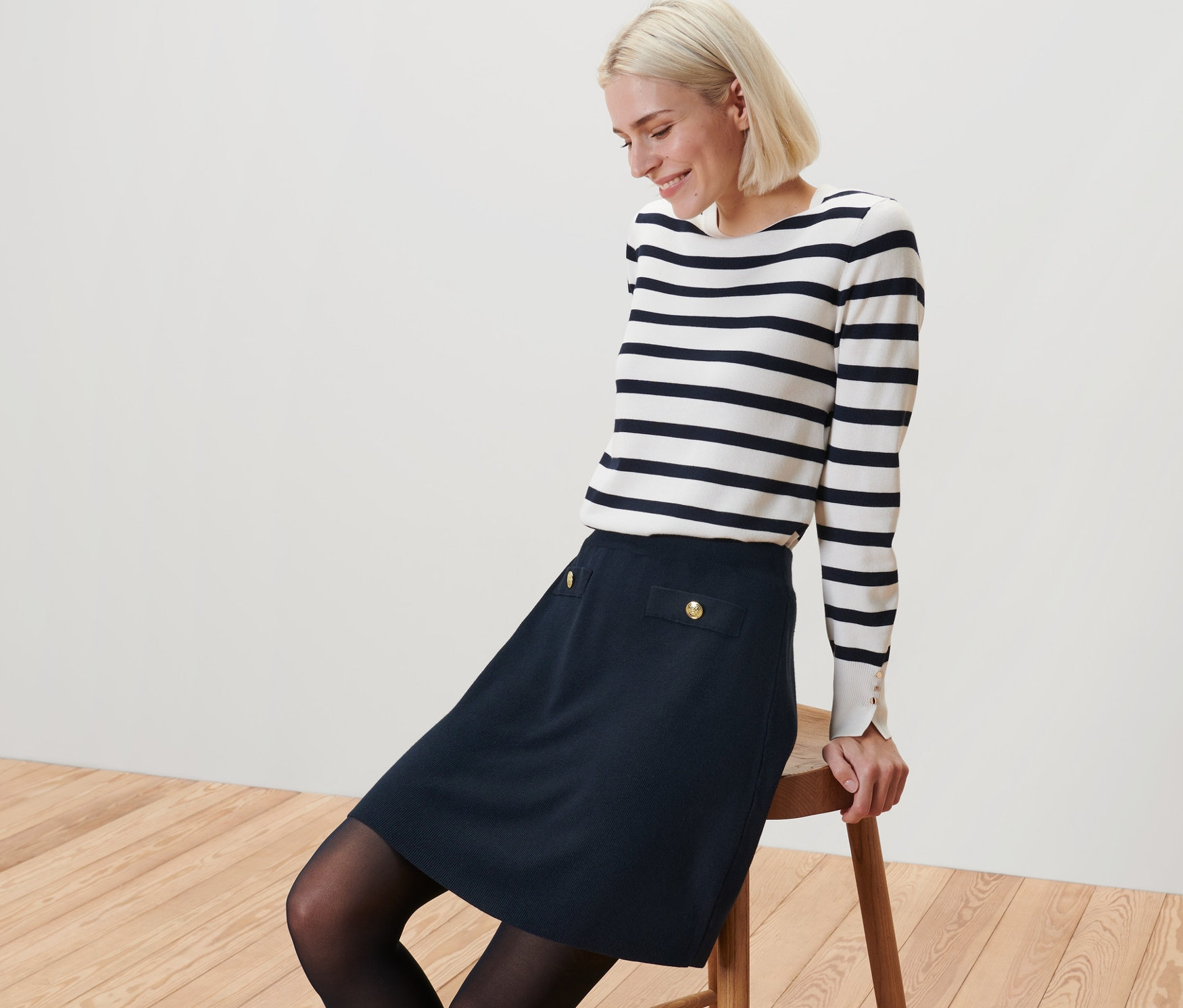 Blondýna sedí na stoličce a má na sobě pletenou sukni s ozdobnými knoflíky a pruhovaný top.