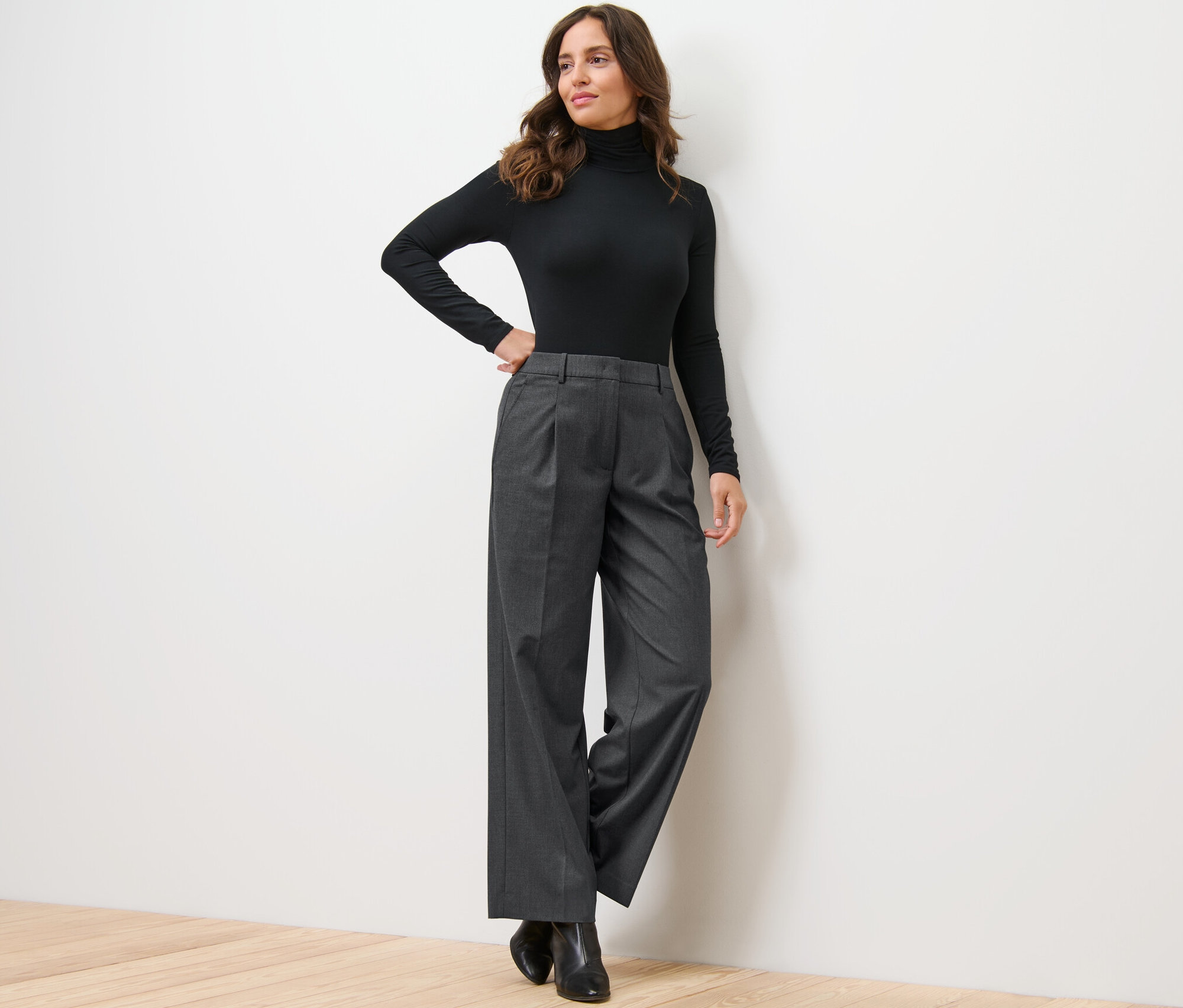 Žena pózuje opřená o stěnu v černém roláku a pohodlných tkaných kalhotách.