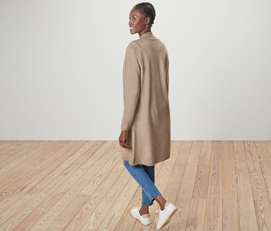 Žena pózuje v otevřeném dlouhém kardiganu, modrých džínách a bílých botách.