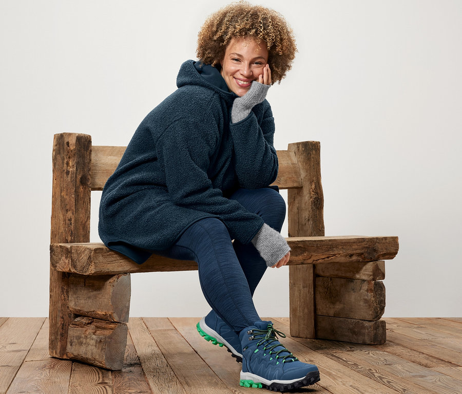 Žena sedí na dřevěné lavici a má na sobě plyšový kabát oversized a sportovní legíny.