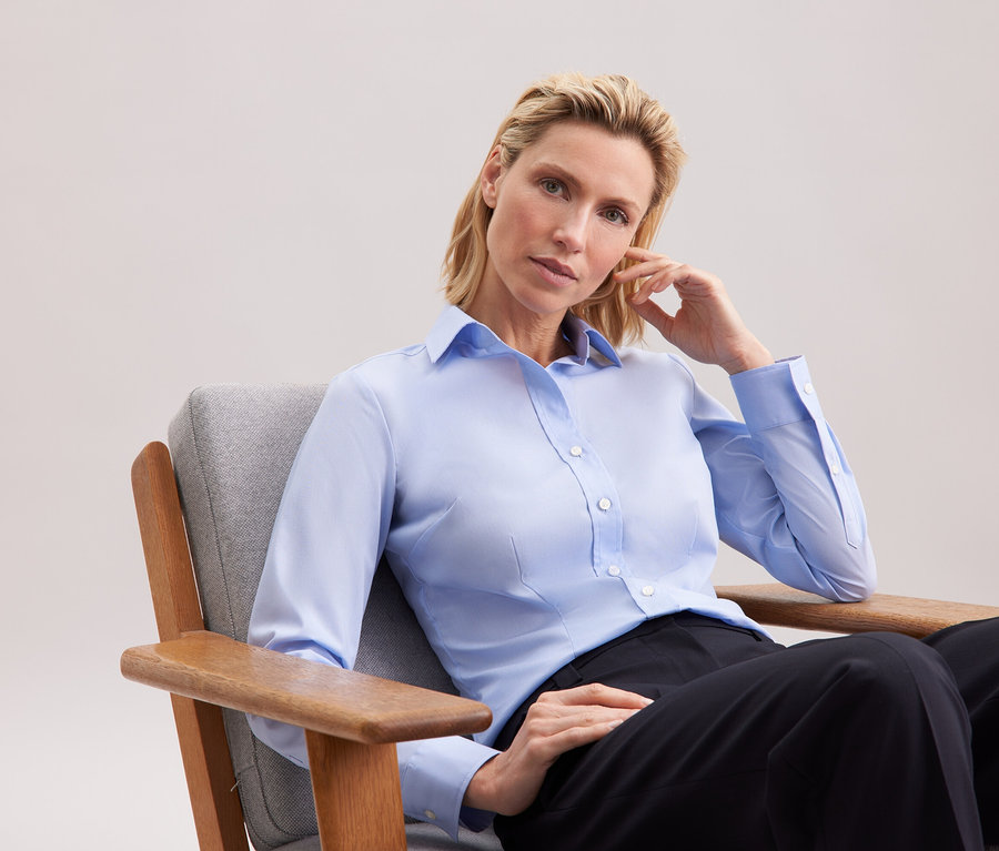 Blondýna sedí v šedém křesle a má na sobě světle modrou košili a černé kalhoty.