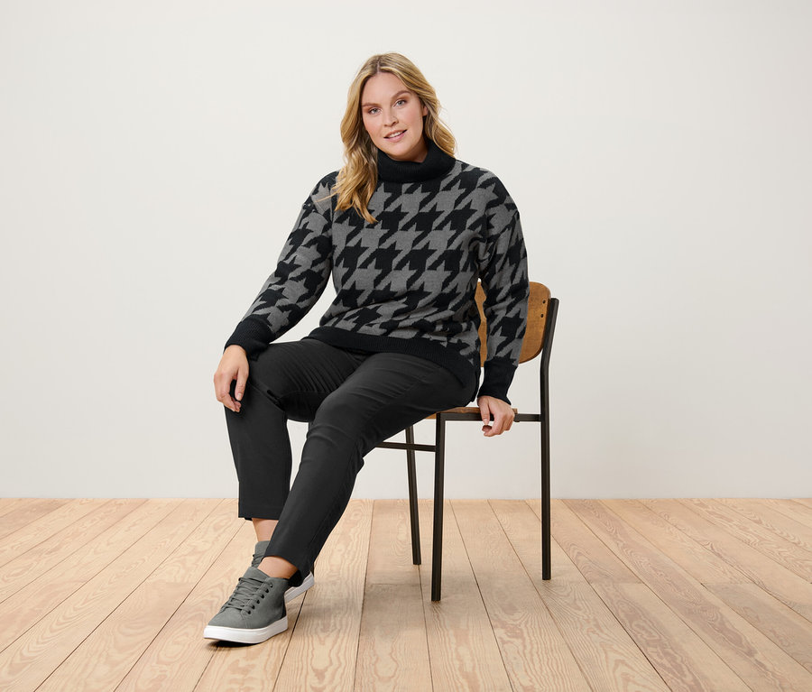 Žena sedí na židli a má na sobě svetr se vzorem kohoutí stopy, černé kalhoty a šedé tenisky.