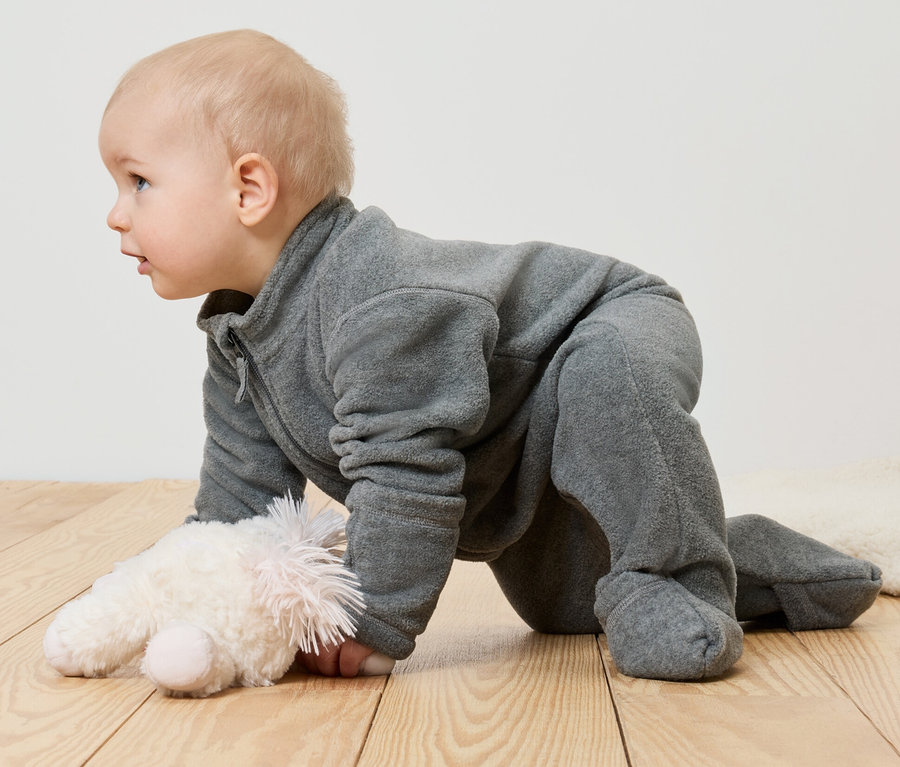 Kojenecký fleecový overal na miminku ležícím na břiše vedle plyšové hračky.