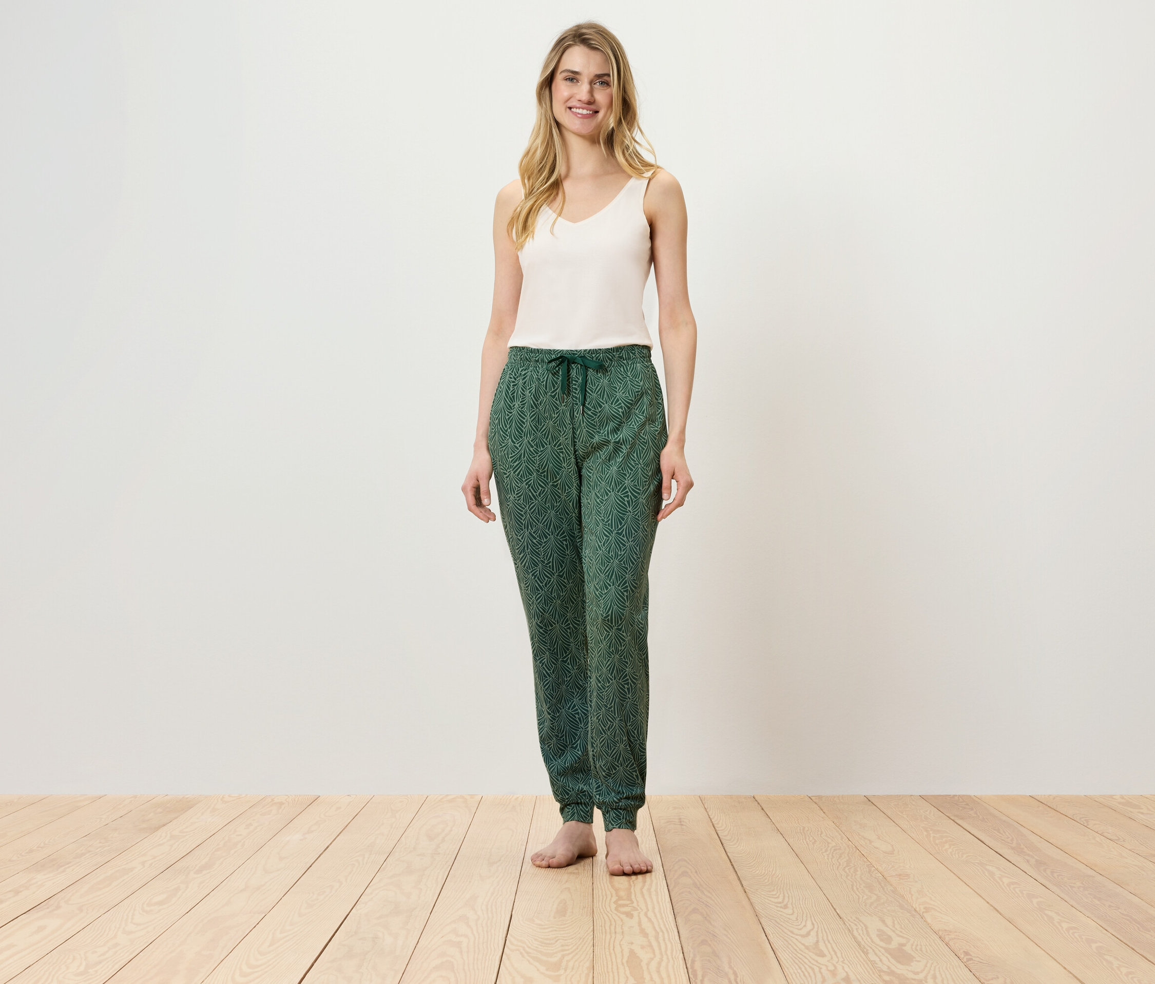 Žena stojí na dřevěné podlaze a má na sobě bílý top a zelené kalhoty.