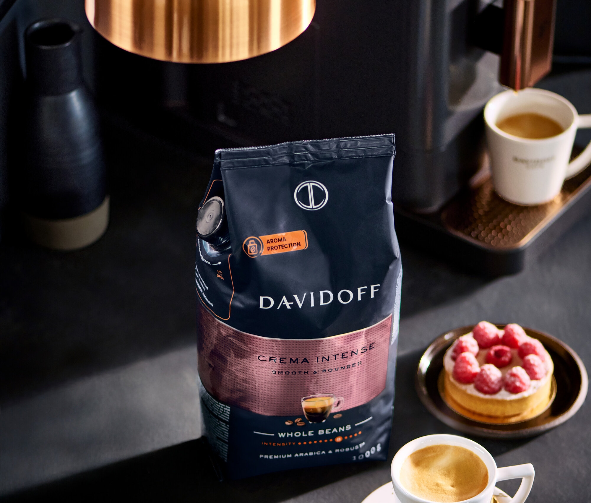 Balíček DAVIDOFF CAFÉ Crema Intense 1000 g zrnkové kávy stojí vedle šálku kávy a malinového koláče.