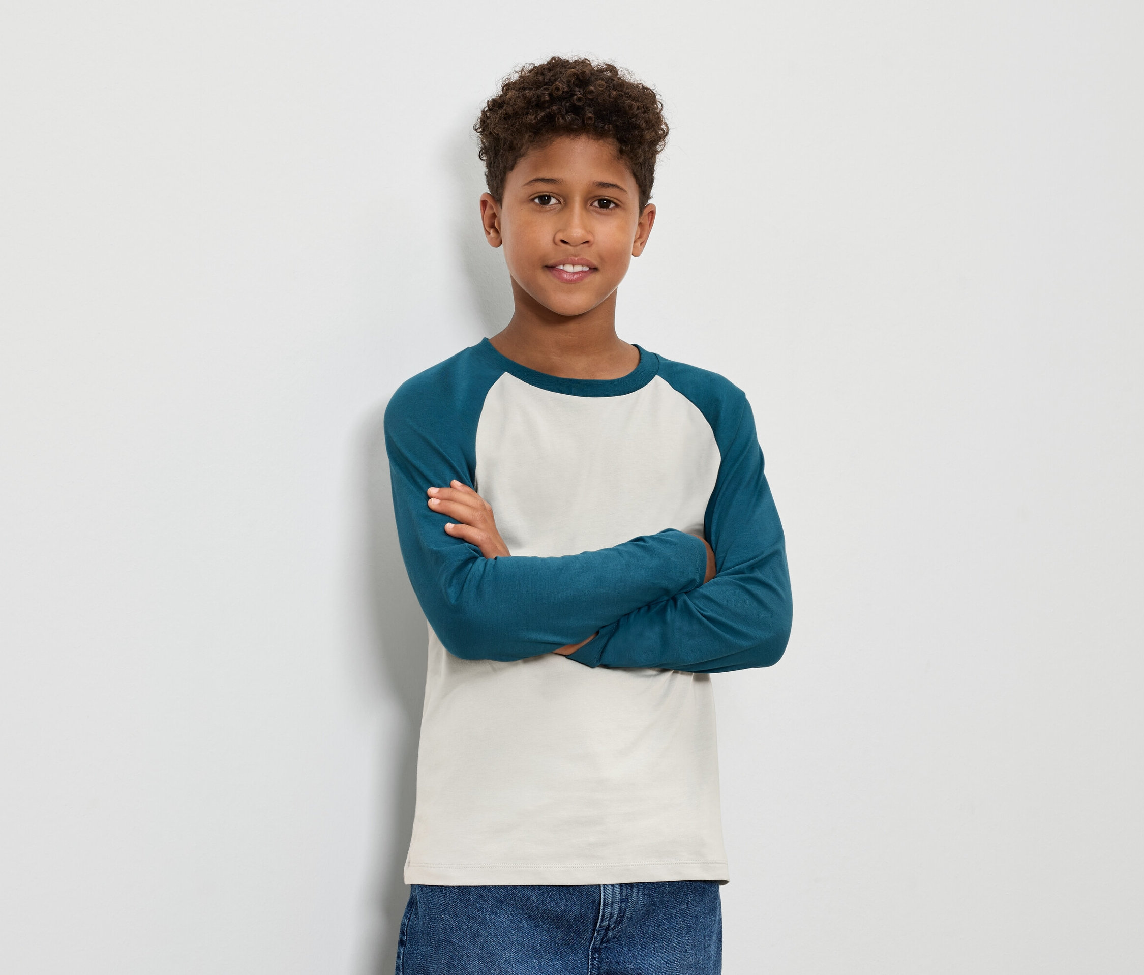 Chlapec s hnědými kudrnatými vlasy stojí s rukama zkříženýma na hrudi a má na sobě modrozelené tričko s dlouhým rukávem a džíny.