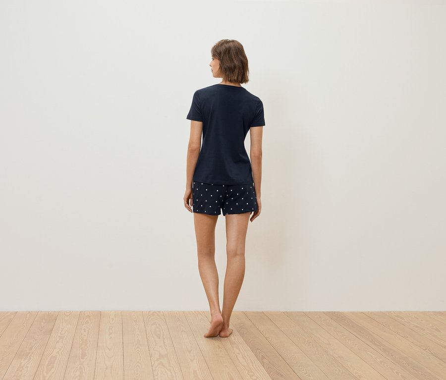 Žena stojí zády k fotoaparátu a má na sobě tmavě modré tričko a šortky s bílými puntíky.