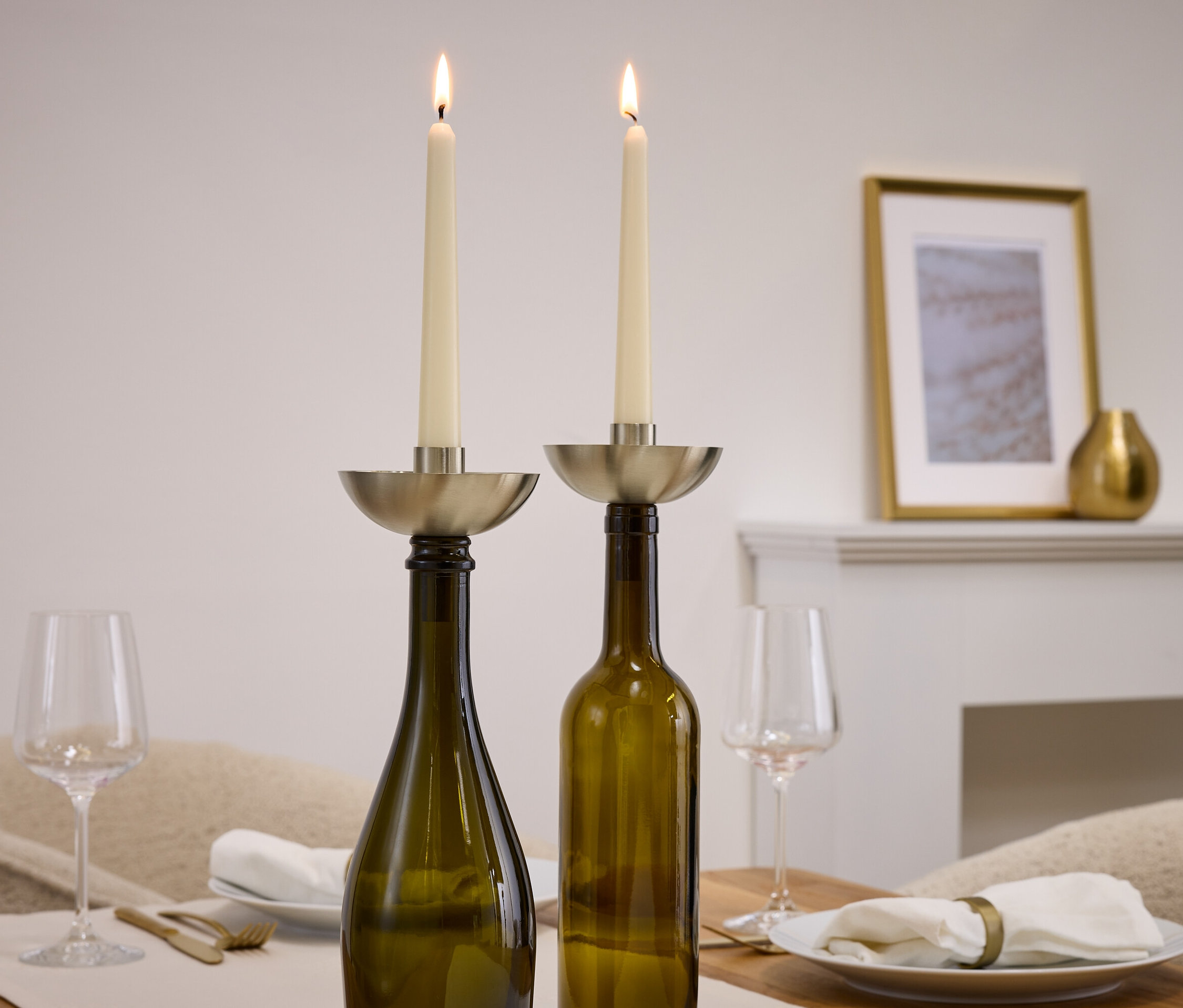 Dvě zapálené svíčky ve stříbrných svícnech umístěných na zelených lahvích, vedle jsou sklenice a talíře.