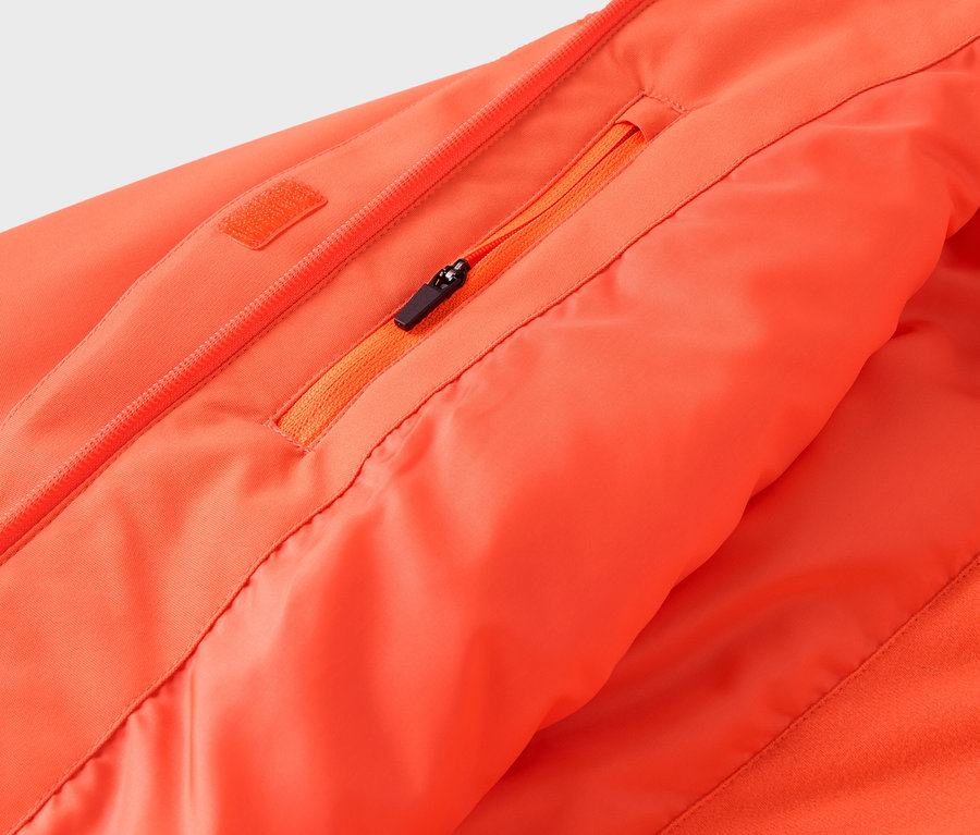 Detail oranžové bundy se zipem a kapsou.