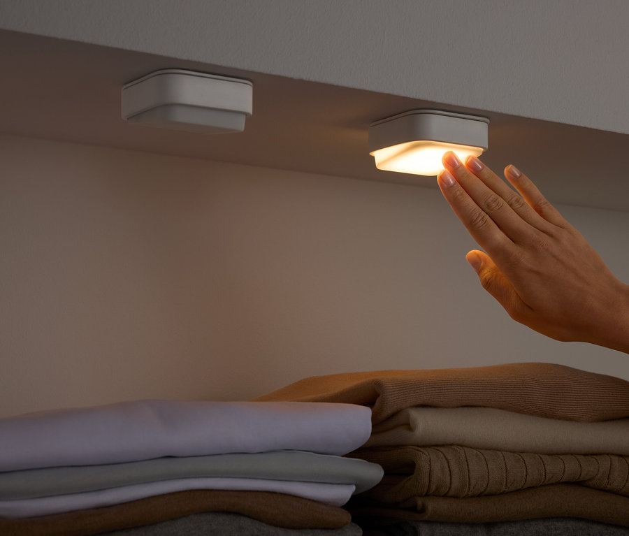 Ruka se dotýká čtvercového světla pod policí nad složeným oblečením.