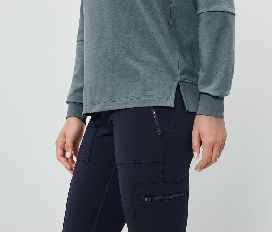 Detail ženy nosící modré kalhoty s kapsami na zip a šedozelený top s dlouhým rukávem.