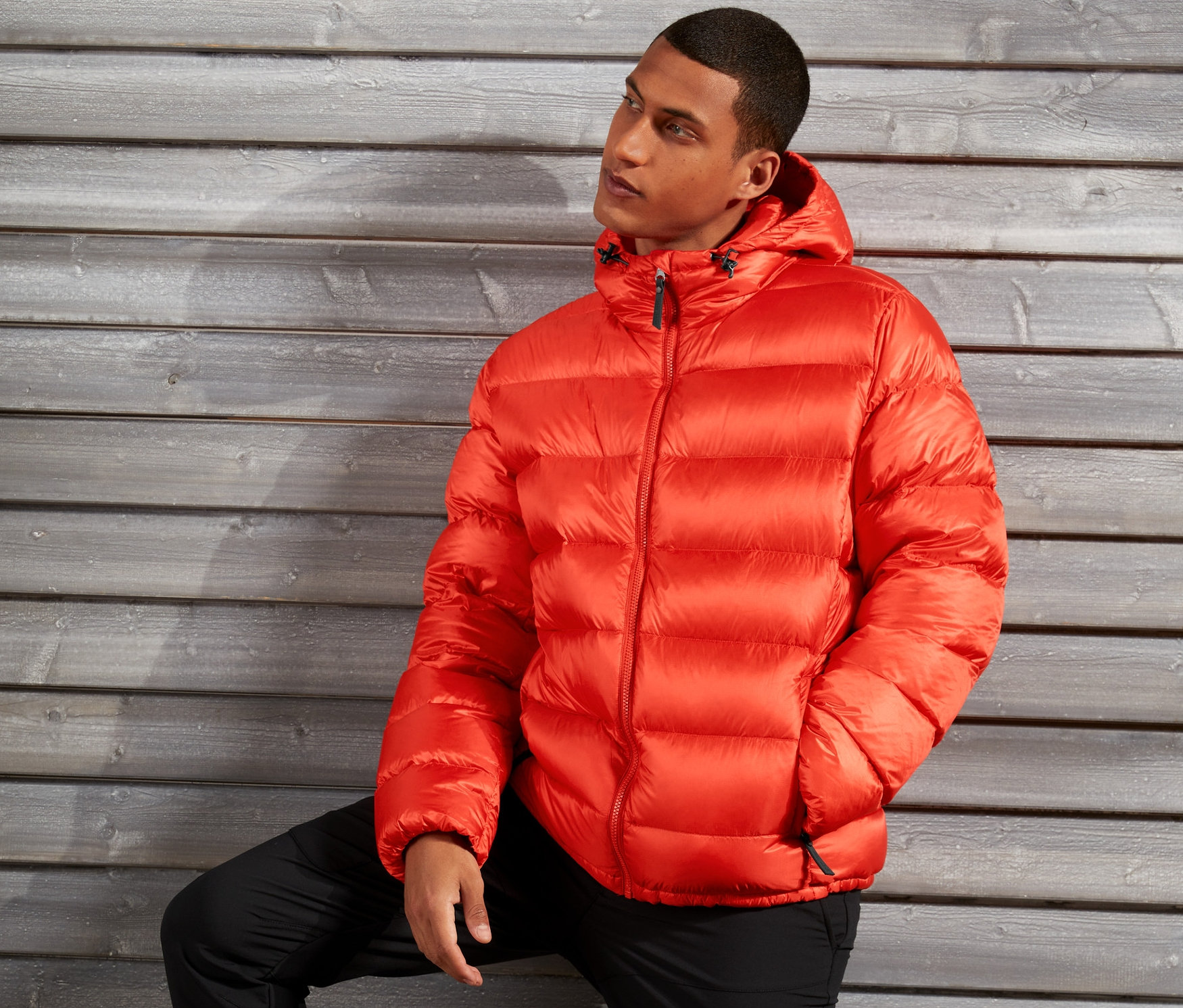 Muž pózuje v oranžové péřové bundě před dřevěnou stěnou.