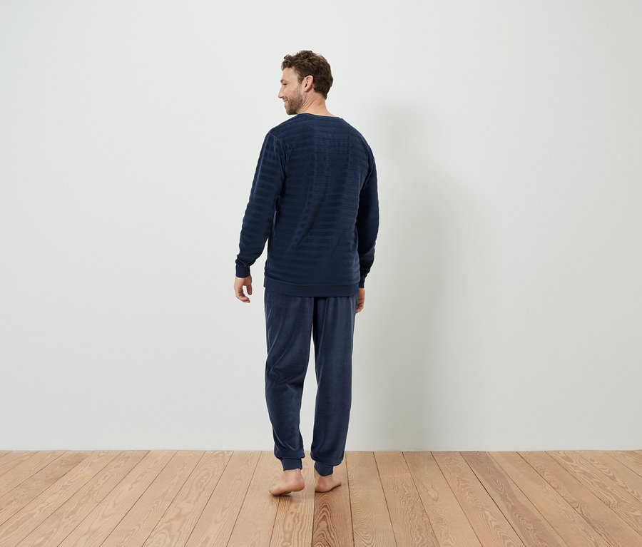 Muž v tmavě modrém pruhovaném pyžamu stojí zády ke kameře na dřevěné podlaze.