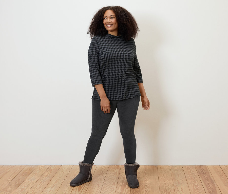 Žena pózuje v černých manšestrových legínách, vzorovaném topu a kozačkách.