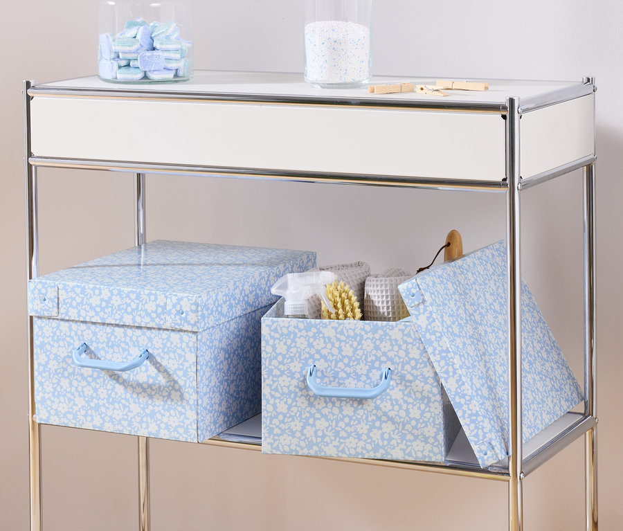 Na polici stojí dva modré úložné boxy s víkem a úložný box s víkem uvnitř s bílými ručníky a kartáčem.