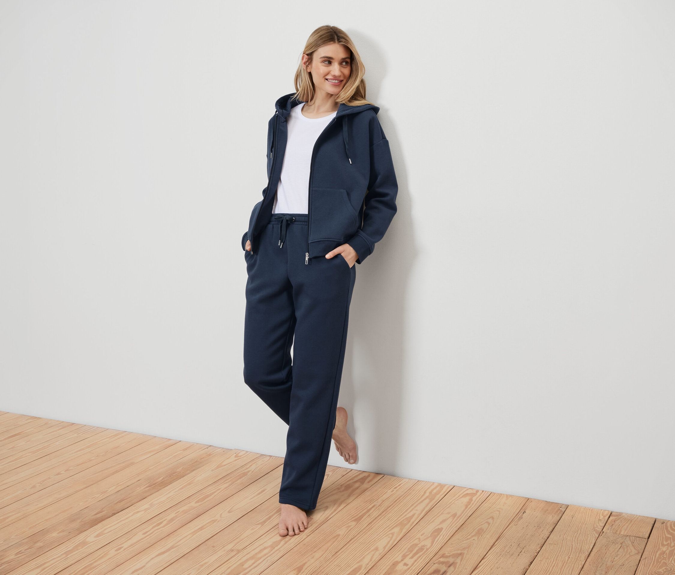 Žena v tmavě modré teplákové soupravě opřená o bílou zeď.