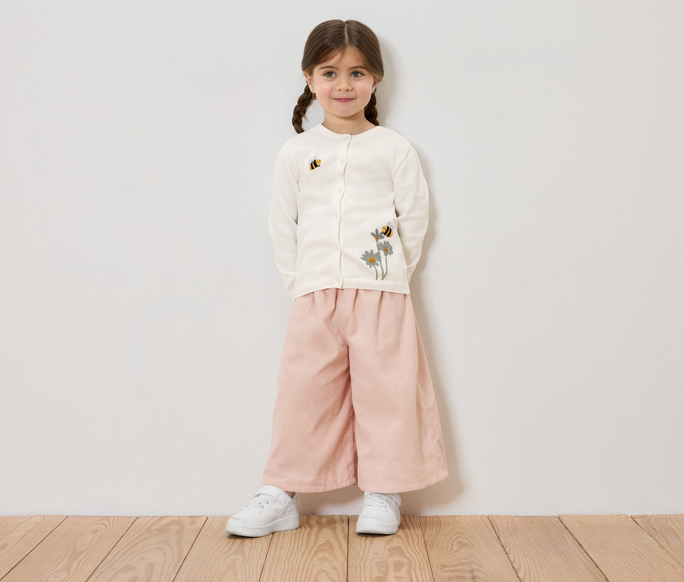 Dívka s copánky má na sobě krémový kardigan s květinovou a včelí výšivkou, růžové kalhoty a bílé tenisky, na pozadí bílé zdi.