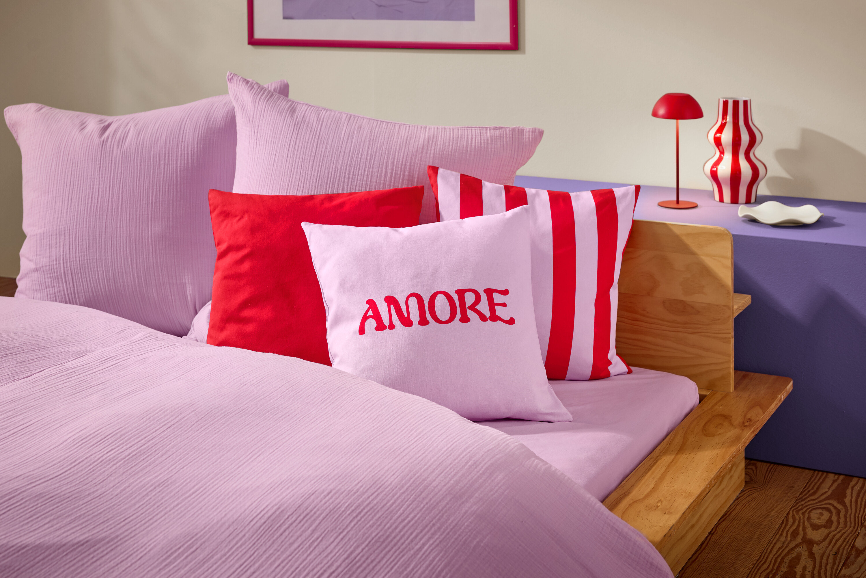 Detailní záběr postele s růžovým ložním prádlem a polštáři. Na jednom z polštářů je červeným písmem napsáno slovo AMORE.