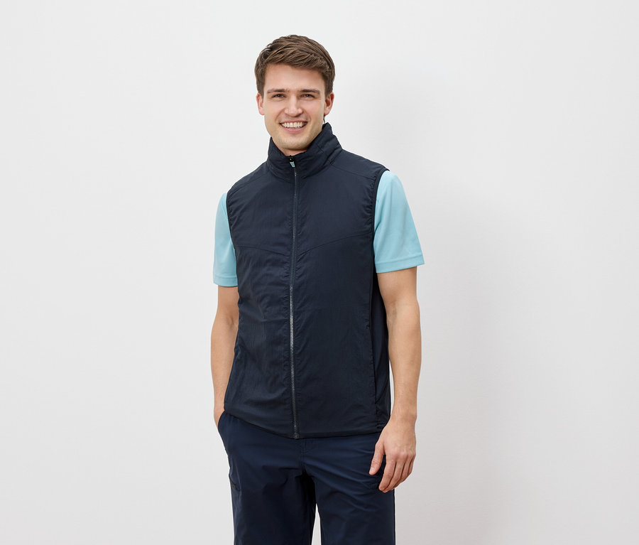 Muž se usmívá do kamery a má na sobě tmavě modrou vestu přes světle modré tričko.