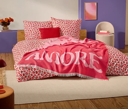 Na posteli s červeno-bílým leopardím vzorem leží červená deka s nápisem "AMORE" velkými bílými písmeny.