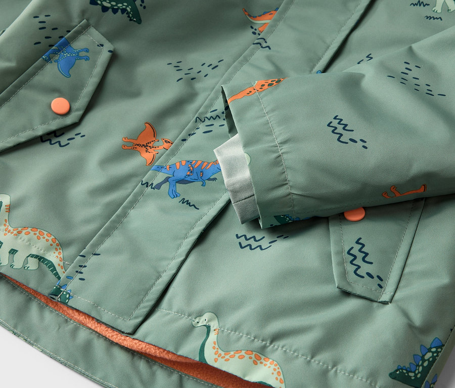 Detailní záběr světle zelené bundy se vzorem dinosaurů, zapínáním na knoflíky a kapsami.