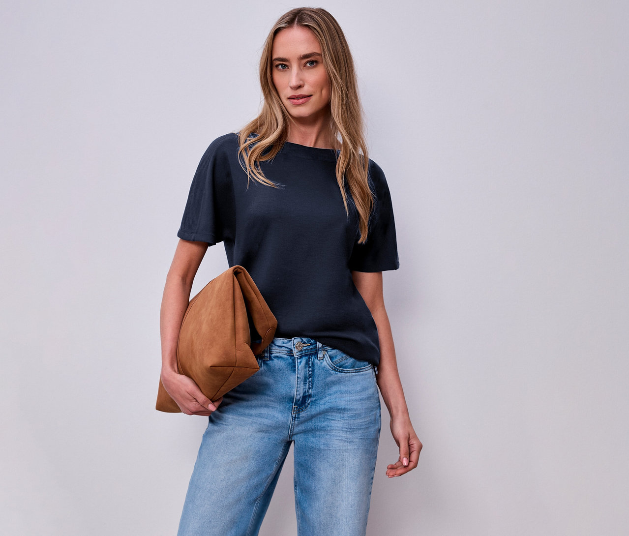 Žena s hnědou taškou a tričkem s krátkým rukávem Street One, námořnická modrá, před bílou stěnou.