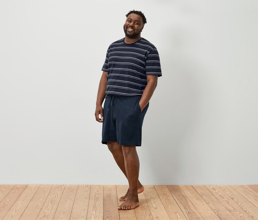 Usmívající se muž stojí bosý před bílou stěnou a má na sobě pruhované tričko a tmavě modré šortky.