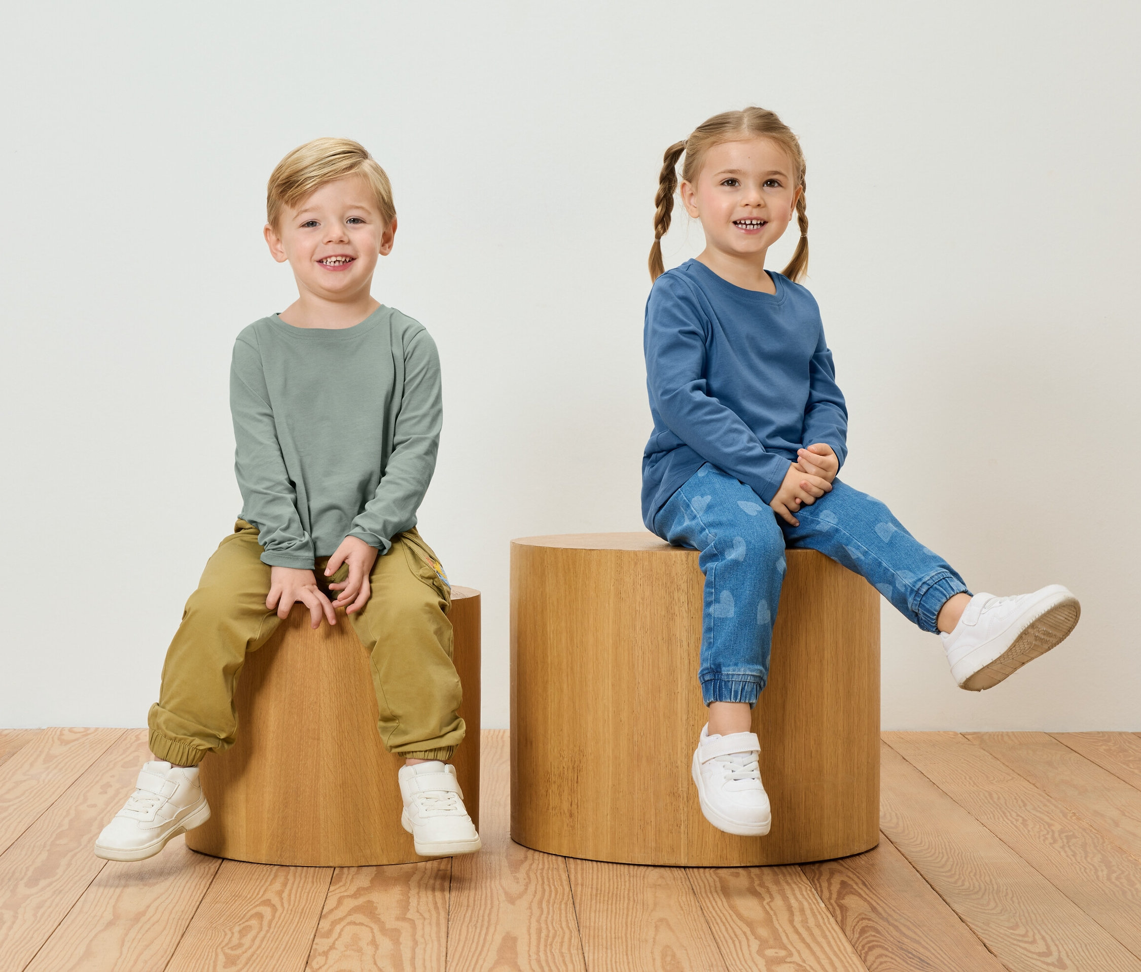 Chlapec v zeleném tričku s dlouhým rukávem a khaki kalhotách sedí vedle dívky v modrém tričku s dlouhým rukávem a džínách se vzorem srdce na dřevěných stoličkách.