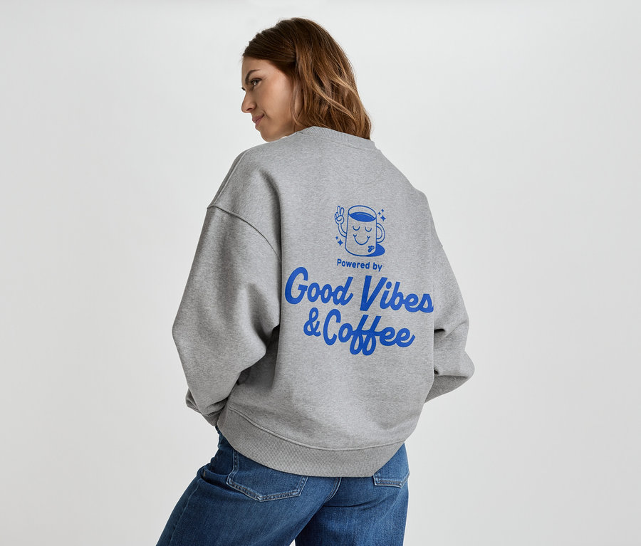 Žena má na sobě šedou mikinu s modrým nápisem "Good Vibes & Coffee" na zádech a džíny.
