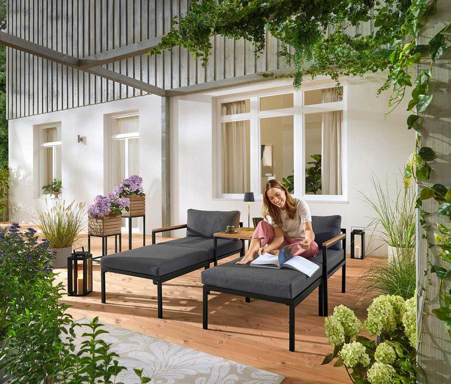 Žena sedí na šedém zahradním lehátku a čte si knihu. V pozadí jsou vidět dva sloupky na rostliny „Elin“.