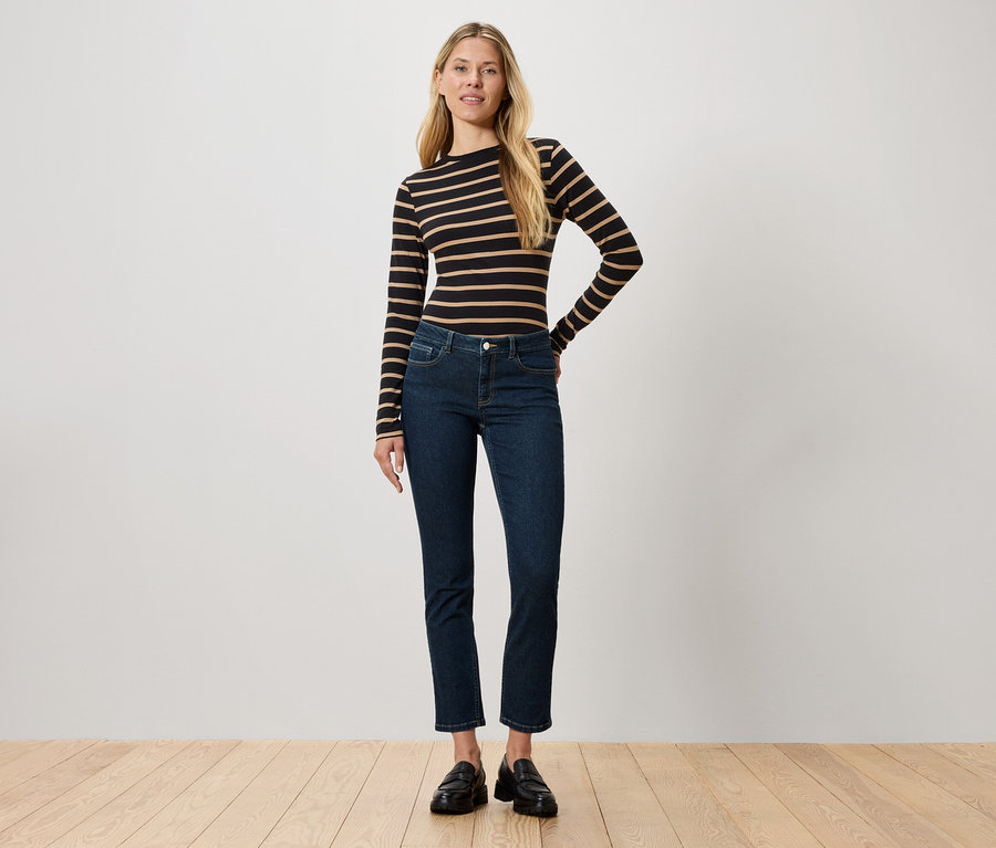 Žena stojí a má na sobě černo-hnědé pruhované tričko s dlouhým rukávem, tmavé džíny a černé boty.
