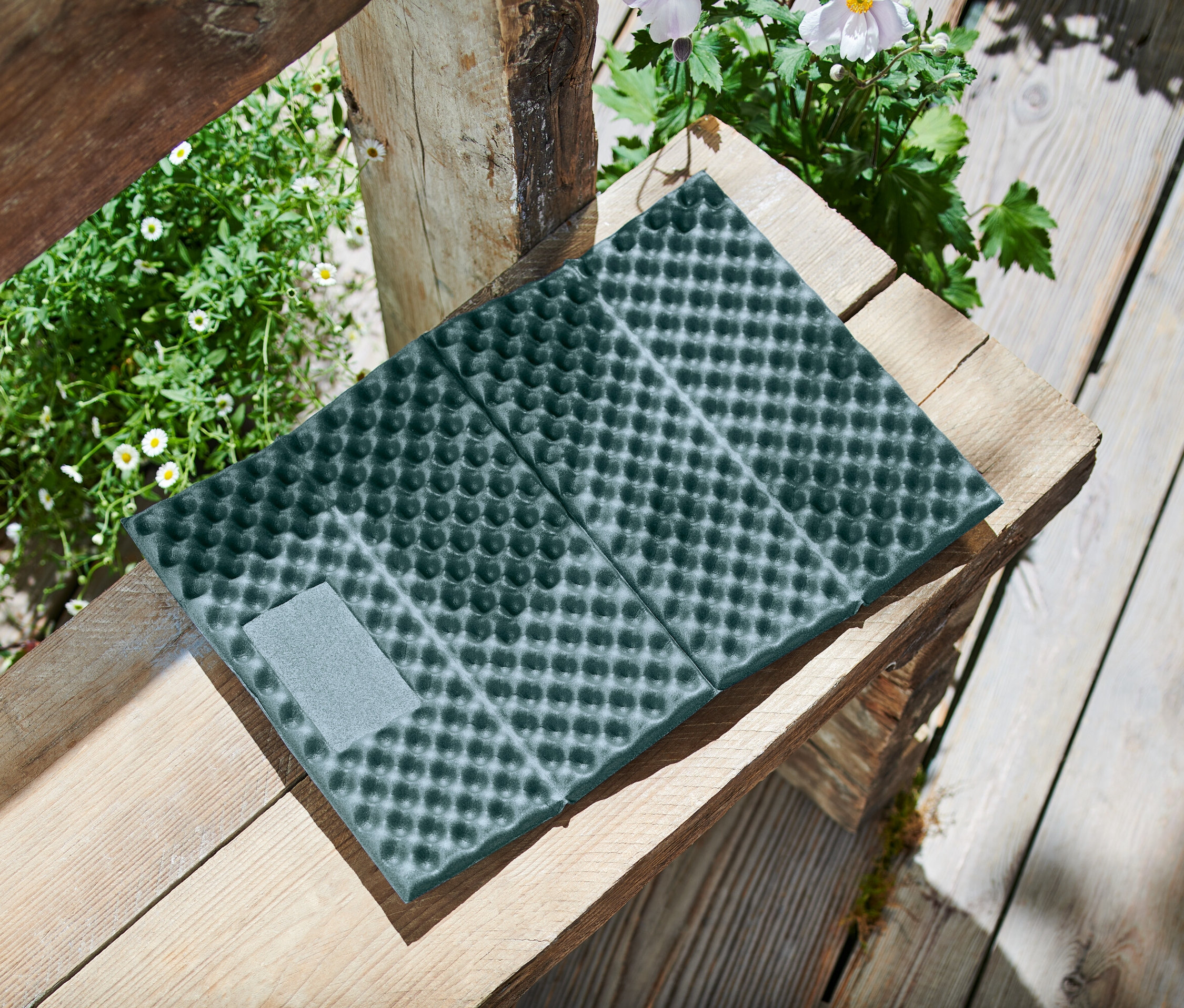 Na dřevěné lavičce leží složená, tmavě zelená podložka na sezení s tečkovaným vzorem a šedým suchým zipem, obklopená rostlinami.
