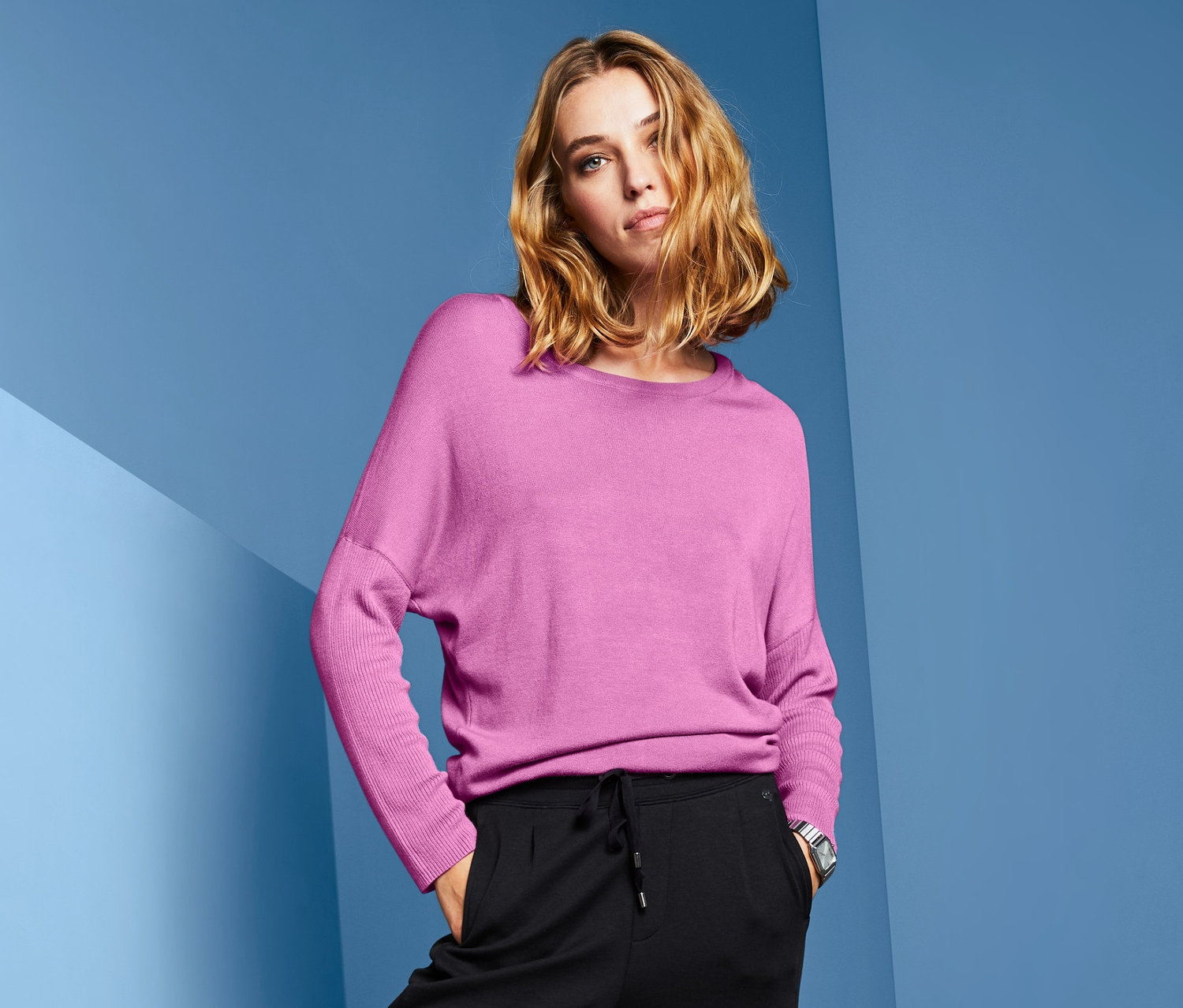Žena pózuje v růžovém svetru z jemné pleteniny s netopýřími rukávy, černých kalhotách a stříbrných botách.