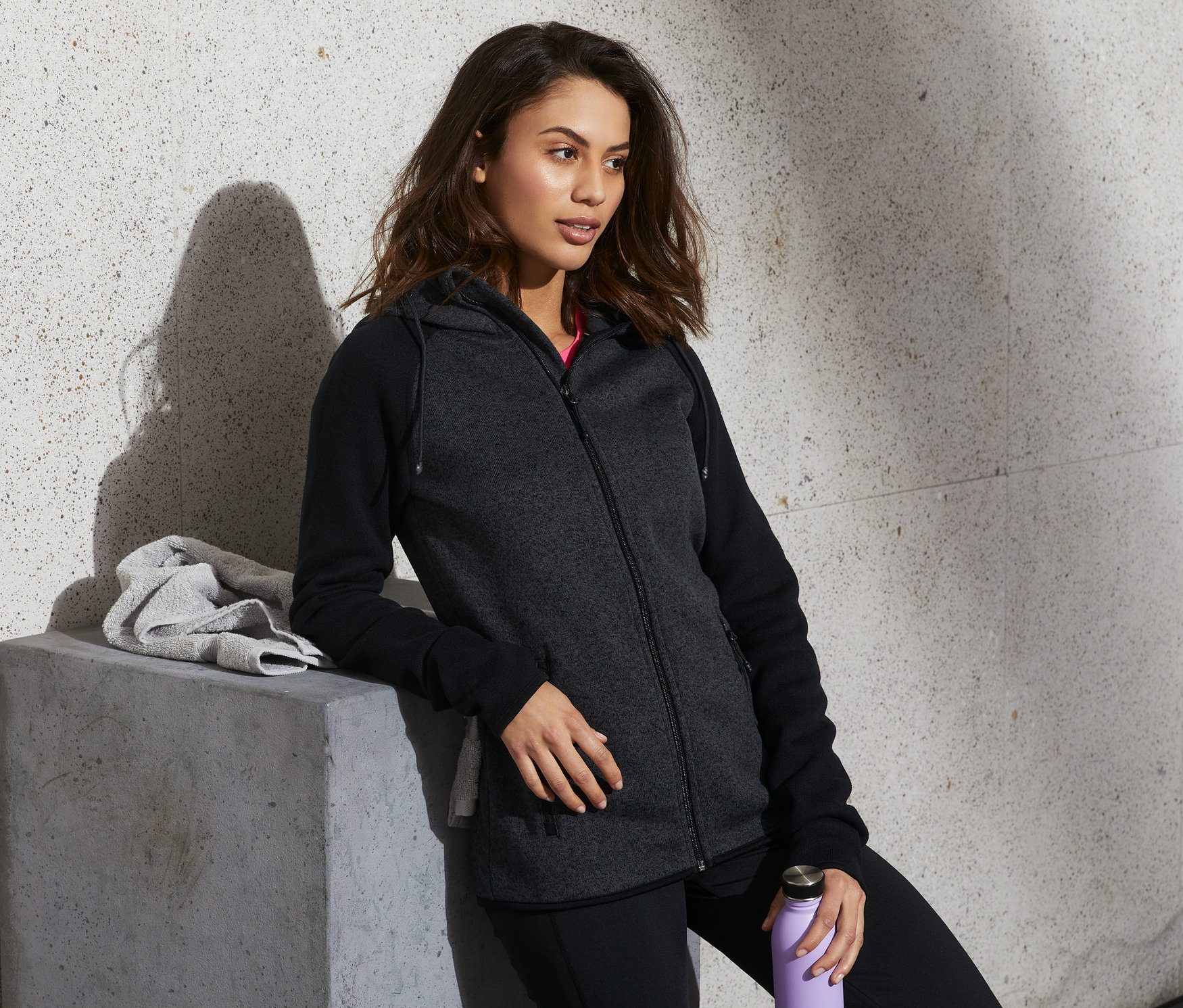 Žena opřená o betonový blok má na sobě mikinu z pleteného fleecu, černé kalhoty a drží fialovou láhev s vodou.