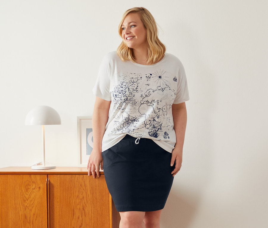 Žena stojí vedle skříňky a má na sobě bílé tričko s kulatým výstřihem a sukni z teplákoviny.