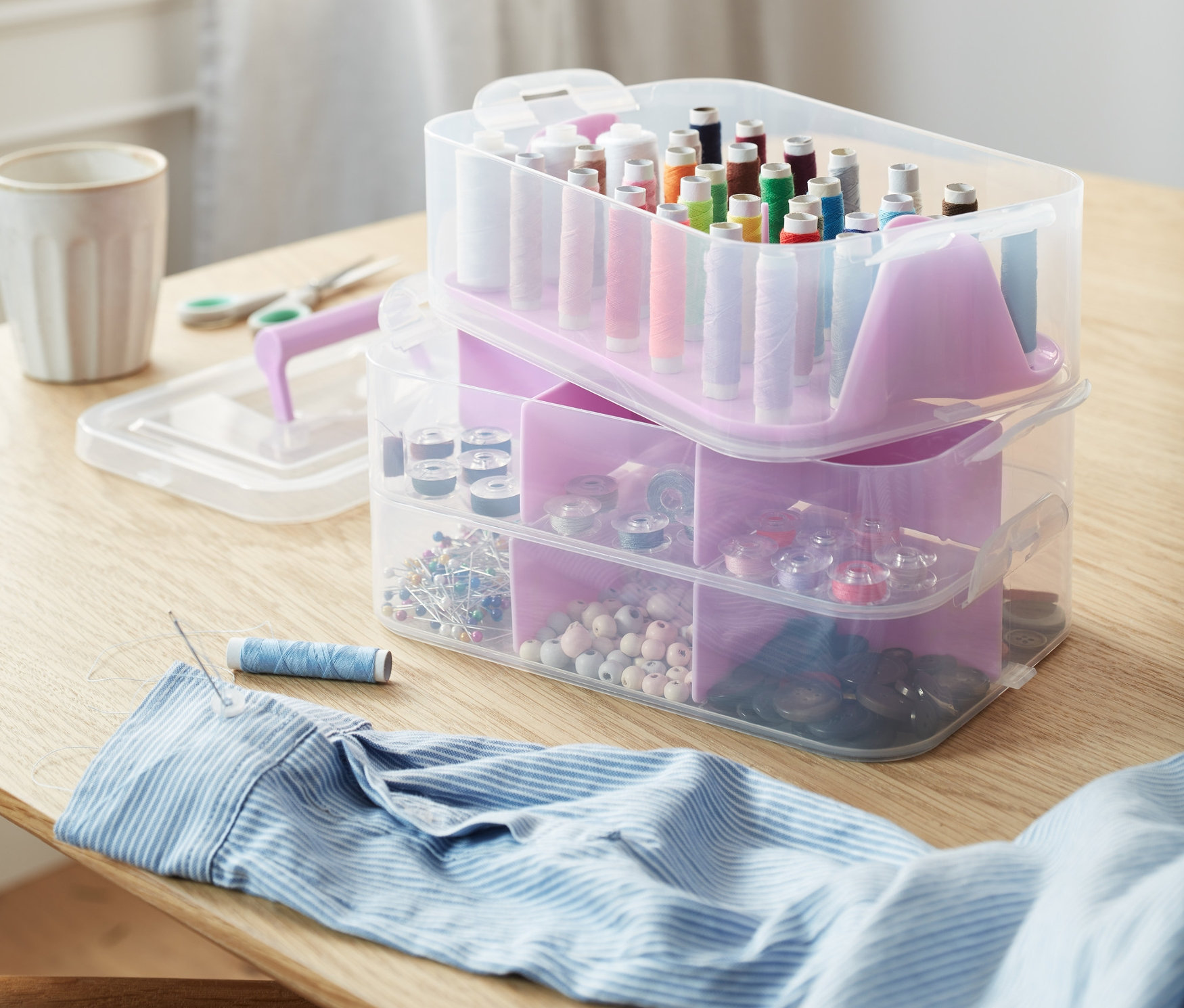 Na stole jsou úložné boxy, nůžky, knoflíky, špulky nití a jehla s nití v košili.