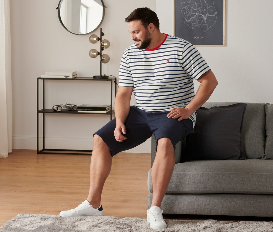 Muž sedí na pohovce a má na sobě boty sneaker, chino šortky v námořnické modré barvě a proužkované tričko s kulatým výstřihem (2 ks).