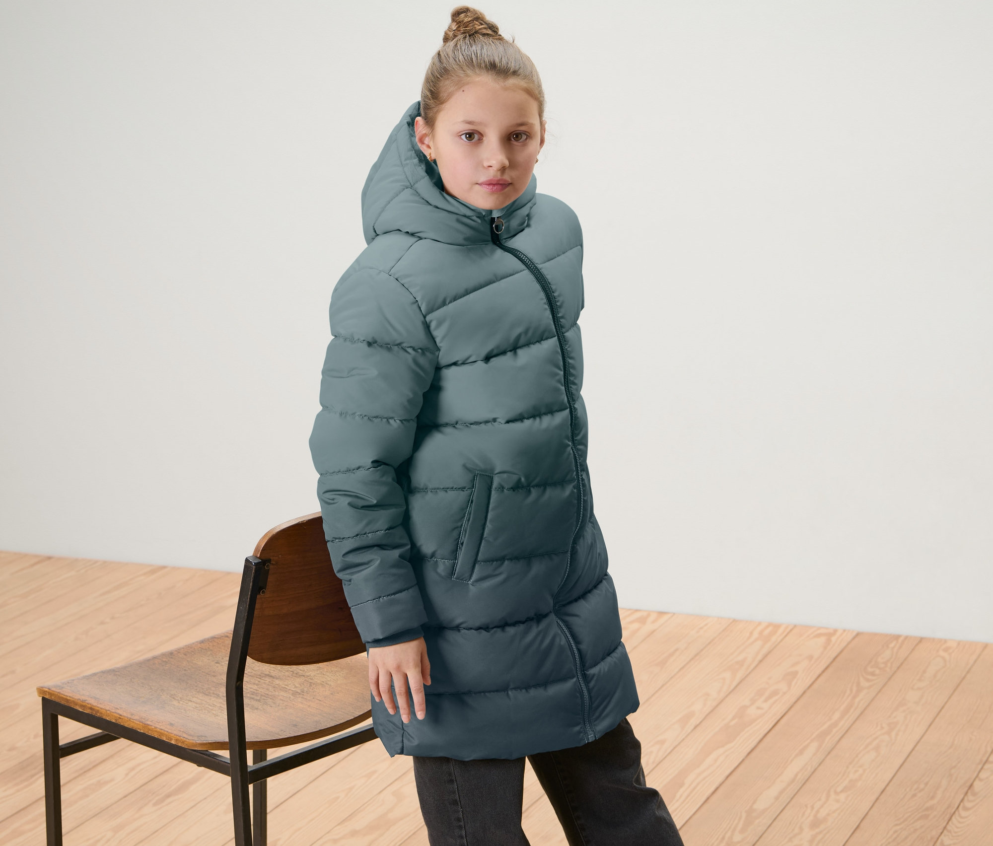 Dívka má na sobě dětský termokabát s barevným přechodem a stojí vedle dřevěné židle.